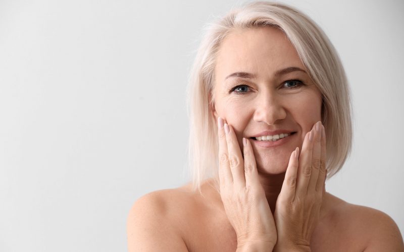 Mature woman giving herself face massage on light background aged-women-face-massage